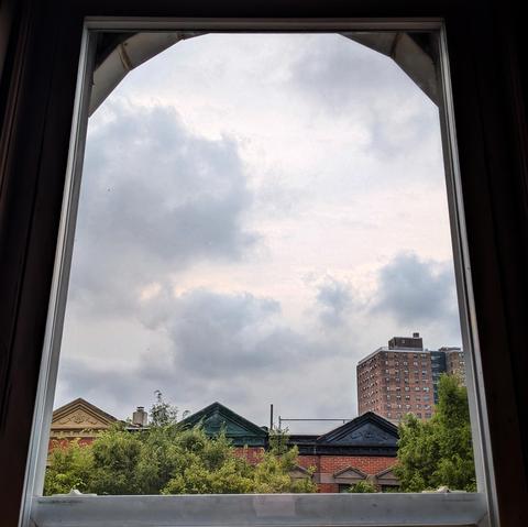 Looking through an arched window two hours after sunrise the sky is light gray, with large darker gray clouds moving through. Pointed roofs of Harlem brownstones with red brickwork are across the street, and a taller apartment building can be seen in the distance. The green tops of two trees are on the bottom and right.