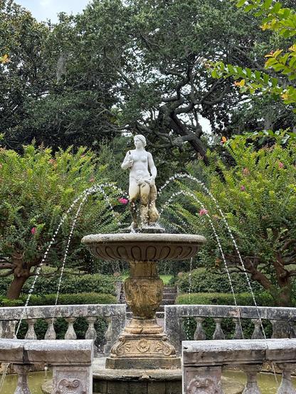 A decorative fountain featuring a statue of a nude woman holding a small object, surrounded by water jets. The fountain is set in a lush garden with various greenery and trees, adding to the serene atmosphere of the scene.