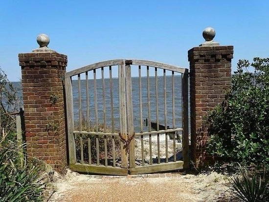 A weathered wooden gate stands between two brick columns, leading to a view of a calm body of water. Surrounding the gate are lush green plants and a clear blue sky.