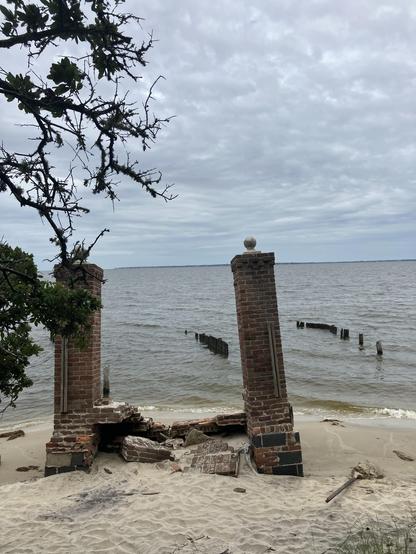 The image shows two crumbling brick columns on a sandy shore, with a view of a body of water and a cloudy sky. The remnants of wooden posts are visible in the water, indicating the outline of a structure. A sparse tree branches overhead.