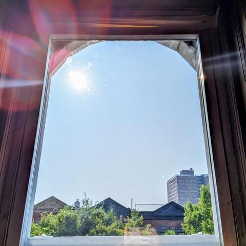 Looking through an arched window three hours after sunrise the sun is above the buildings in the cloudless light-blue sky, making a dramatic lens flare with multi-colored petals in the upper left. Pointed roofs of Harlem brownstones are silhouetted across the street, and a taller apartment building can be seen in the distance. The green tops of two trees are on the bottom and right.