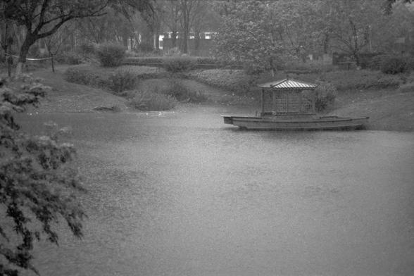 Ilford Pan 400 (FF)

**English:**
A serene black-and-white photograph of a small, traditional boat with a covered structure, gently floating on a calm pond. The boat is situated near the edge of the water, surrounded by lush trees and foliage. The scene is tranquil, with a backdrop of tall trees and faint outlines of buildings in the distance, suggesting an urban park setting.

**Chinese:**
一张宁静的黑白照片，展示了一艘带有遮盖结构的传统小船，轻轻漂浮在平静的池塘上。小船位于水边，周围环绕着茂密的树木和植被。场景宁静祥和，背景是高大的树木和远处建筑物的淡淡轮廓，暗示这是一个城市公园的环境。