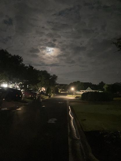 A dimly lit residential street at night, featuring a cloudy sky with a partially visible moon. Streetlights illuminate sections of the road, while vehicles are parked along the sides. Wet pavement reflects light, adding to the ambiance.