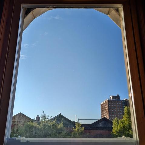 Looking through an arched window and hour after sunrise the deep blue sky is marked with a few tiny wisps of white cloud. Pointed roofs of Harlem brownstones are silhouetted across the street, and a taller apartment building can be seen in the distance. The green tops of two trees are on the bottom and right.