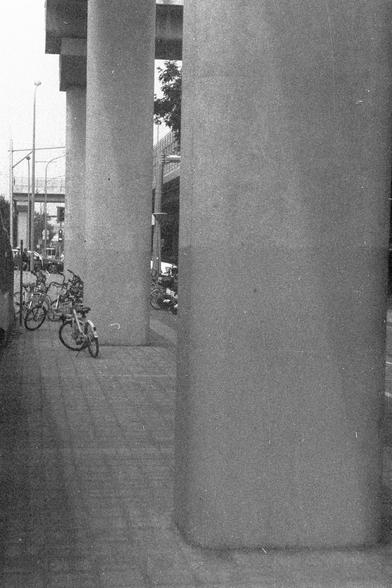 Ilford FP4 Plus 125 (FF)

**English:**
A black and white photograph taken from a narrow alleyway or corridor between large concrete pillars. The perspective is looking out towards a street where several bicycles are parked. The scene captures an urban environment with a mix of architectural elements and everyday life.

**Chinese:**
一张黑白照片，拍摄于大型混凝土柱之间的狭窄小巷或走廊。视角朝向一条街道，那里停放着几辆自行车。这张照片捕捉了一个城市环境，混合了建筑元素和日常生活。