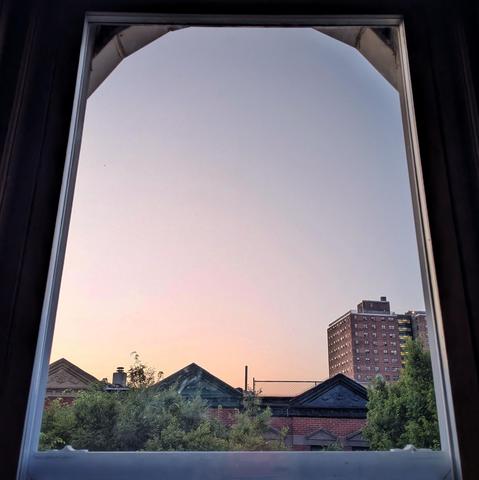 Looking through an arched window seven minutes before sunrise the cloudless sky fades from pale orange at the horizon to light dusty blue up above. Pointed roofs of Harlem brownstones are silhouetted across the street, and a taller apartment building can be seen in the distance. The green tops of two trees are on the bottom and right.
