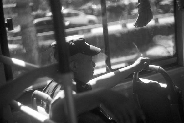 Ilford FP4 Plus 125 (FF)

English Alt Text:
Black and white photo of a bus interior with a person seated at the wheel. Their arm is extended, gripping the steering wheel. Outside the window, blurred cars and greenery suggest motion or a busy roadside.

中文替代文字：
一张黑白照片，展示公交车内部，一人坐在驾驶座，伸出手握住方向盘。窗外模糊的车辆和绿植暗示车辆正在行驶或停在繁忙路边。