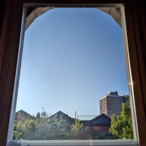Looking through an arched window an hour after sunrise the cloudless sky fades from very light blue at the horizon to pale blue up above. Pointed roofs of Harlem brownstones are silhouetted across the street, and a taller apartment building can be seen in the distance. The green tops of two trees are on the bottom and right.