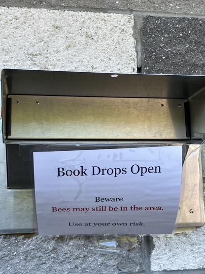 A metal book drop with an open slot is mounted on a textured wall. A sign below warns that "Bees may still be in the area," and advises users to "Use at your own risk."