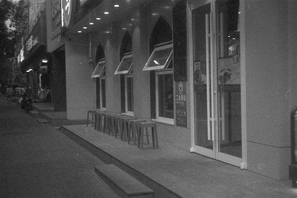Ilford FP4 Plus 125 (FF)

• English Alt Text: The image shows the exterior of a building with a row of stools lined up outside. The building has several arched windows with awnings, and there is a glass door on the right side. There are signs with text in Chinese on the wall next to the door. The scene appears to be on a quiet street with other buildings visible in the background.
• Chinese Alt Text: 这张图片展示了一栋建筑的外部，外面排着一排凳子。建筑有几个带遮阳篷的拱形窗户，右侧有一扇玻璃门。门旁的墙上有一些中文标志。场景似乎在一条安静的街道上，背景中可以看到其他建筑。