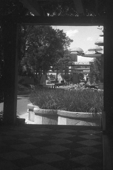 Ilford FP4 Plus 125 (FF)

• English Alt Text: A black-and-white photo of an outdoor urban space, framed by a dark structure in the foreground. The scene includes a large planter with tall grass, a checkered pavement, trees, pedestrians, a car, and dome-shaped architectural structures in the background.
• Chinese Alt Text: 一张黑白照片，展示了一个户外城市空间，前景由一个深色结构框住。画面中有一个种满高草的大型花盆、棋盘格铺地、树木、行人、一辆汽车，以及背景中的圆顶建筑和独特设计的楼房。