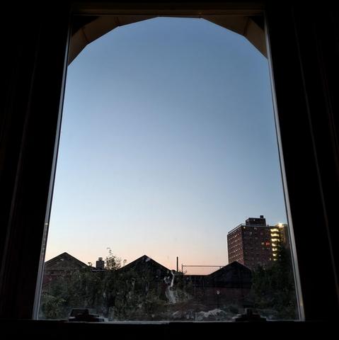 Looking through an arched window half an hour before sunrise the cloudless sky fades from pale peach at the horizon to powder blue up above. Pointed roofs of Harlem brownstones are silhouetted across the street, and a taller apartment building can be seen in the distance.