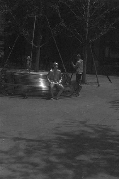 Ilford FP4 Plus 125 (FF)

• English alt text: A circular bench made of stacked metal rings surrounds a tree in an urban public space. One person sits on the bench while another stands nearby, possibly taking a photo. The ground is paved, and more trees are visible in the background.
• 中文替代文字：在城市公共空间中，一棵树被由金属环堆叠而成的圆形长椅环绕。一人坐在长椅上，另一人站在旁边，似乎在拍照。地面为铺装路面，背景中还有其他树木