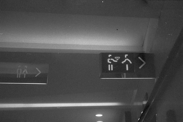 Ilford FP4 Plus 125 (FF)

• English alt text: Two illuminated signs mounted on a ceiling, both with arrows pointing to the right. The left sign shows two stick figures, one holding an object like a bag. The right sign shows two stick figures shaking hands with a diamond shape between them, suggesting a transaction or exchange. The image is in black and white.
• 中文替代文字：两个安装在天花板上的发光指示牌，箭头均指向右侧。左侧标志显示两个火柴人，其中一个拿着类似包的物品。右侧标志显示两个火柴人握手，中间有一个菱形图案，可能表示交易或交换。图像为黑白色调。