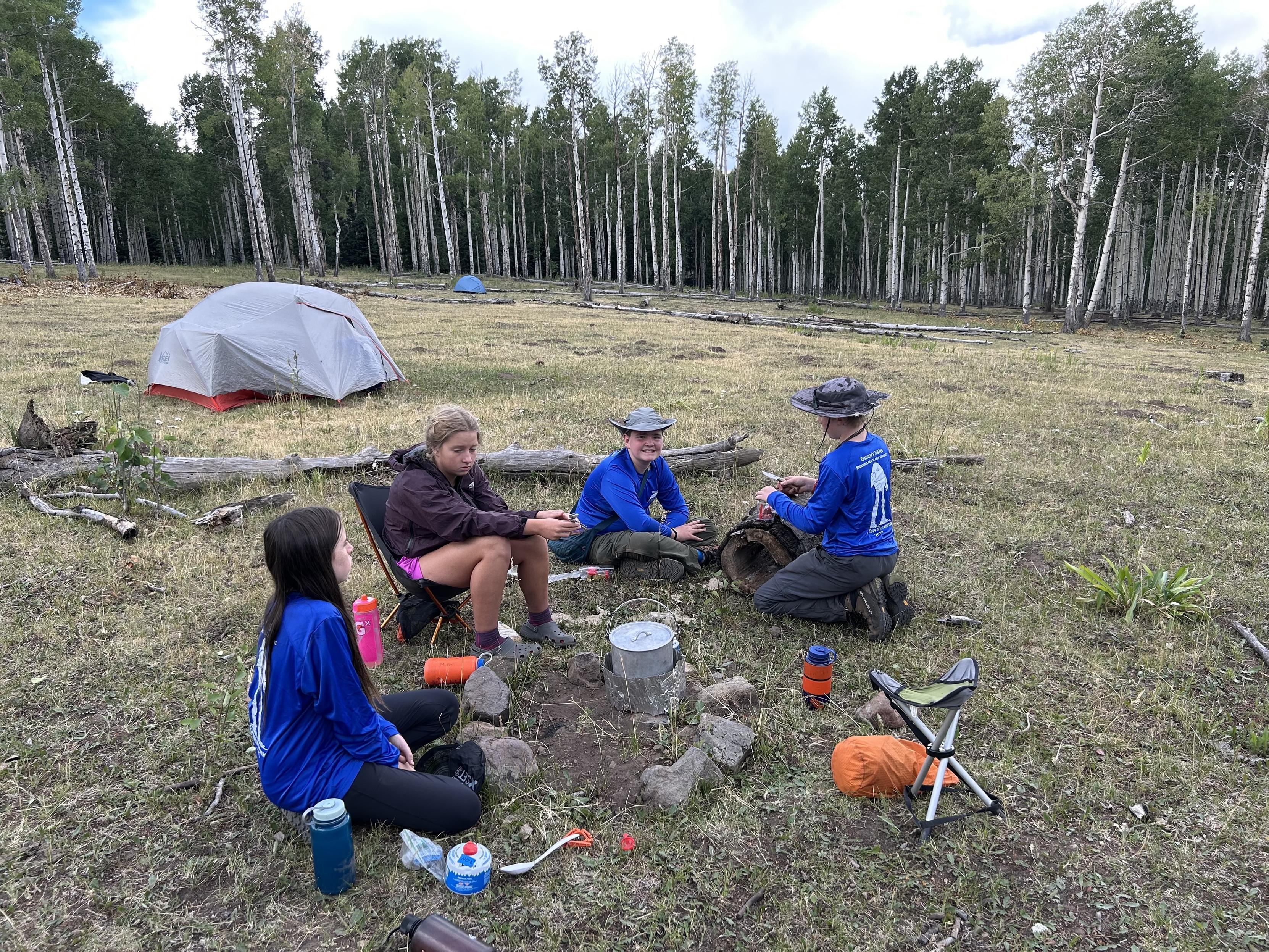 A group of four children is sitting around a campsite with tents in the background. They are engaged in a cooking activity near a small fire pit, surrounded by grass and trees. The atmosphere is relaxed, with the children wearing outdoor clothing and hats.