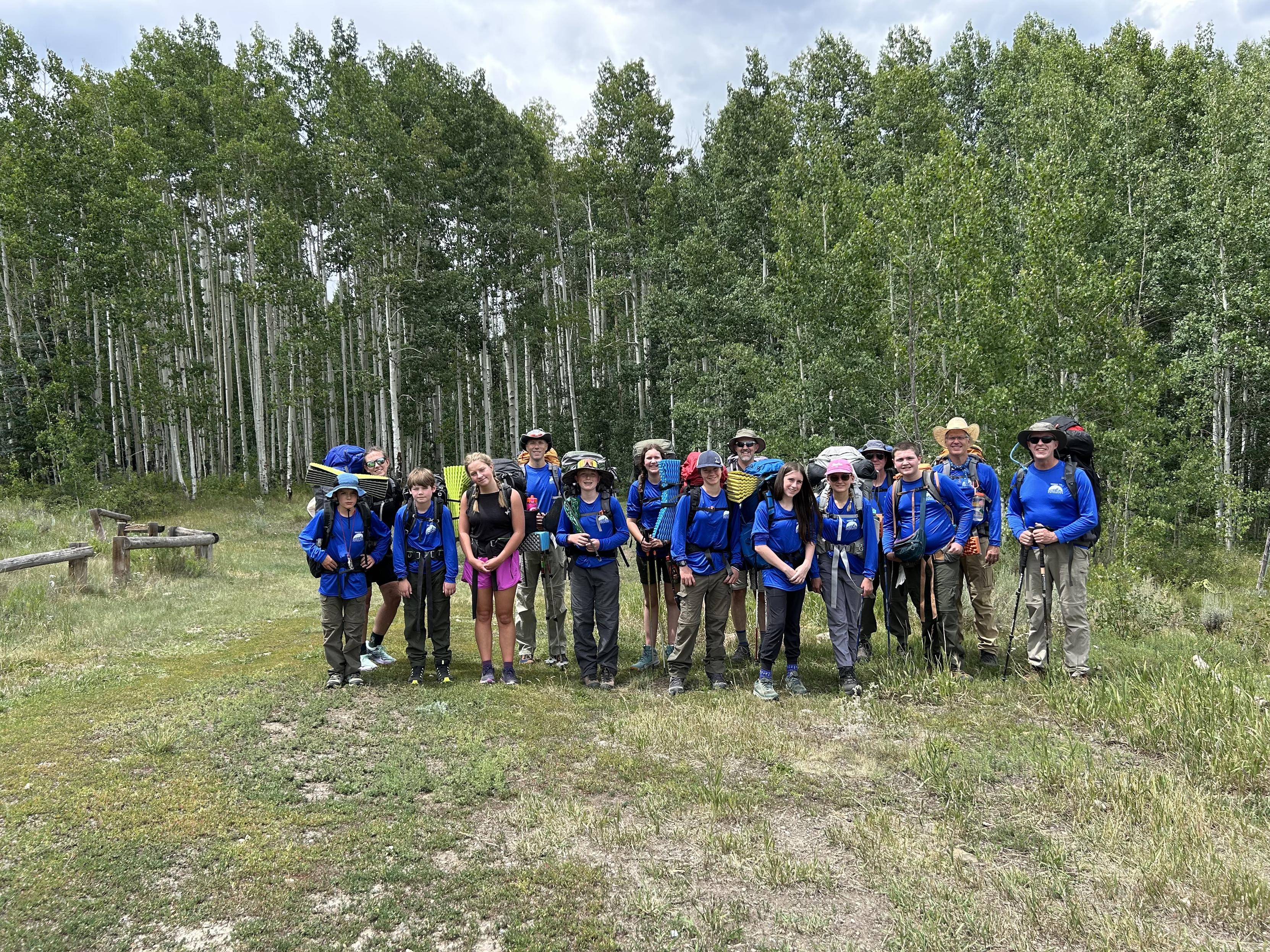 A group of 16 people, including children and adults, stands together in a forested area. They are wearing blue shirts and carrying backpacks, ready for a hiking adventure. The background features tall trees and a grassy clearing.