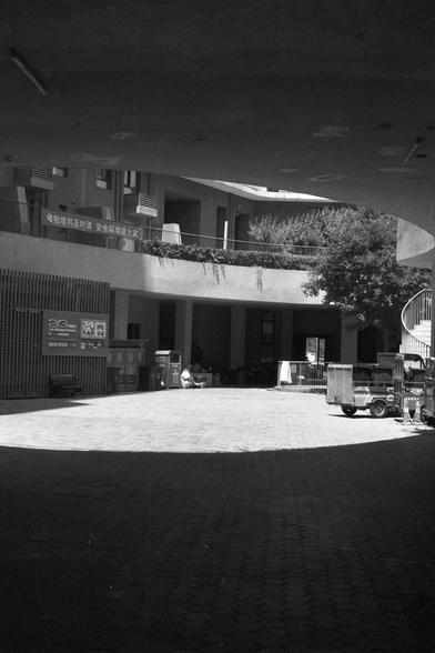 Ilford FP4 Plus 125 (FF)

**English:**
A black-and-white photograph depicts the entrance of a modern building. The entrance is framed by a curved overhang and a large tree in the center of the courtyard. Above the entrance, there is a sign with text, and the building features large windows and a clean, minimalist design. The shadows and light create a dramatic effect, emphasizing the architectural lines.

**Chinese:**
这张黑白照片展示了一座现代建筑的入口。入口处由一个弧形遮阳和庭院中央的一棵大树构成。入口上方有一个带文字的标志，建筑物以大窗户和简洁的极简设计为特色。光影…