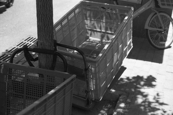Ilford FP4 Plus 125 (FF)
**English:**
A black-and-white image shows a close-up of two plastic crates stacked on a handcart. The crates are placed on a sidewalk next to a tree and a bicycle. The shadows of the tree leaves are cast on the ground, adding texture and contrast to the scene.

**Chinese:**
这张黑白照片展示了两个塑料箱叠放在手推车上的特写。箱子放在人行道上，旁边是一棵树和一辆自行车。树叶的影子投射在地面上，为场景增添了纹理和对比。