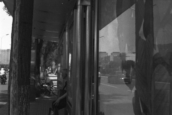 Ilford FP4 Plus 125 (FF)
**English:**
A black-and-white photograph captures a moment at a bus stop or shelter. The scene is framed by large vertical metal poles and glass panels. Outside the shelter, a person is seated on a bench, partially visible through the glass. The reflection on the glass reveals a street scene with vehicles and buildings, adding depth and a sense of urban life to the composition.

**Chinese:**
这张黑白照片拍摄的是一个公交车站或避雨亭。画面由几根大型金属立柱和玻璃面板构成。透过玻璃可以看到亭外有一个人坐在长椅上，身影若隐若现。玻璃的反光映射出街道上…