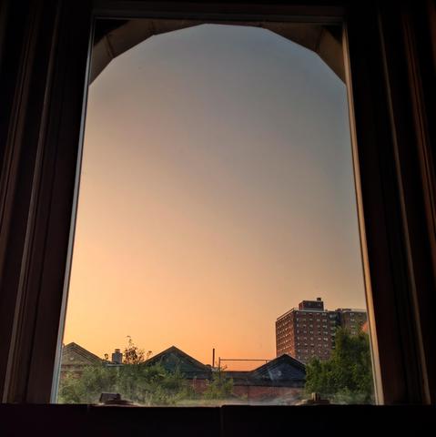 Looking through an arched window fourteen minutes after sunrise the cloudless sky fades from peach at the horizon to steel blue up above. Pointed roofs of Harlem brownstones are silhouetted across the street, and a taller apartment building can be seen in the distance. The green tops of two trees are on the bottom and right. 