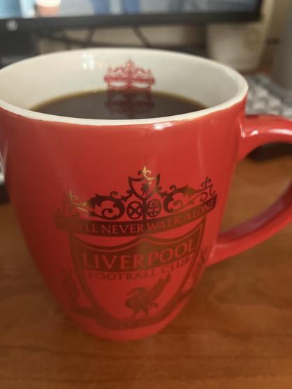 A red mug displaying the Liverpool Football Club logo and the phrase "You'll Never Walk Alone," filled with black coffee, is placed on a wooden surface. In the background, a computer monitor is partially visible.
