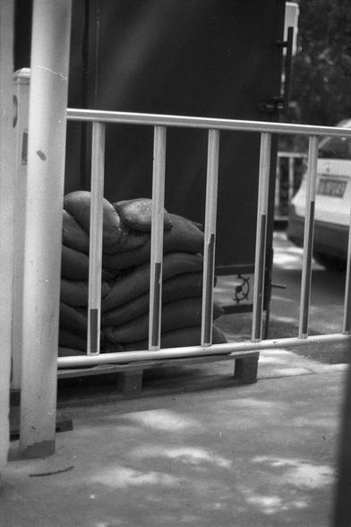 Ilford FP4 Plus 125 (FF)
English Alt Text:
A black and white image showing a stack of sandbags placed on a wooden pallet behind a metal railing. Part of a car and other urban structures are visible in the background.

中文替代文字：
一张黑白照片，画面显示一堆沙袋堆放在木托盘上，位于金属栏杆后方。背景中可见部分汽车和其他城市结构。