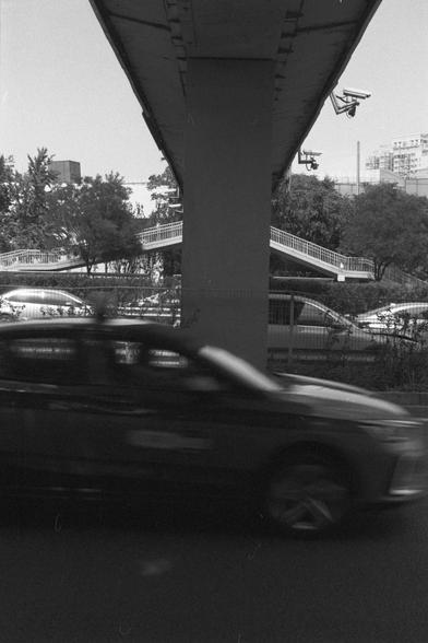 Ilford FP4 Plus 125 (FF)
English Alt Text:
A black and white urban scene featuring a large concrete pillar supporting an overpass. Two surveillance cameras are mounted above. A car speeds past in the foreground, appearing blurred due to motion. Behind it are trees, a pedestrian bridge, parked cars, and a fence.

中文替代文字：
一张黑白城市照片，画面中心是一根支撑高架桥的大型混凝土柱。上方安装了两个监控摄像头。前景中一辆汽车快速驶过，因运动而模糊。背景中有树木、人行天桥、停放的车辆和栅栏。