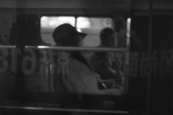 Ilford FP4 Plus 125 (FF)
English Alt Text:
A black and white photo taken from outside a vehicle, likely a bus or train, showing passengers inside. Reflected Chinese text overlays the window, partially obscuring the view. One person wearing a cap and mask is seated in the foreground, while another figure is visible in the dimly lit background. The interplay of shadows and reflections creates a layered, mysterious atmosphere.

中文替代文字：
一张黑白照片，从车外拍摄，画面显示车内乘客，可能是公交车或地铁。窗户上反射出中文文字，部分遮挡了视线。前景中一位戴帽子和口罩…