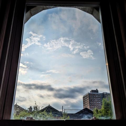 Looking through an arched window two hours after sunrise the blue sky is filled with dark gray clouds above the horizon and swirling white clouds above. Pointed roofs of Harlem brownstones are silhouetted across the street, and a taller apartment building can be seen in the distance. The green tops of two trees are on the bottom and right. 