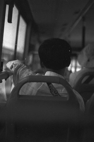 Ilford FP4 Plus 125 (FF)
• English Alt Text: Black and white photo of a person sitting on a bus, viewed from behind. The person wears a light shirt with a strap labeled “FASHION” and rests their arm on the window ledge, gazing outside.
• 中文替代文字： 一张黑白照片，从后方拍摄一位坐在公交车上的人。此人穿着浅色衬衫，肩上背带印有“FASHION”字样，手臂靠在窗台上，望向窗外。