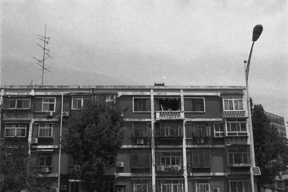 Ilford FP4 Plus 125 (FF)
• English Alt Text: Black and white photo of an old multi-story residential building with balconies, air conditioning units, and a rooftop antenna. A streetlight stands to the right, and trees partially obscure the lower windows.
• 中文替代文字： 一张黑白照片，展示了一栋老旧的多层住宅楼，带有阳台和空调机，屋顶有天线。右侧有一盏路灯，楼前的树木部分遮挡了底层窗户。