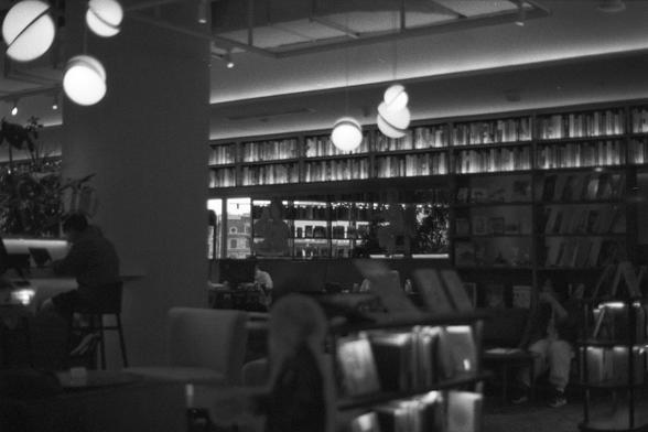 Ilford FP4 Plus 125 (FF)
• English Alt Text: Black and white photo of a quiet library or bookstore interior with bookshelves lining the walls, soft hanging lights, and people seated at tables reading or working. A large column stands in the center, and plants are visible on the left.
• 中文替代文字： 一张黑白照片，展示了一个安静的图书馆或书店内部，墙边排列着书架，吊灯柔和，几位人坐在桌旁阅读或工作。画面中央有一根大柱子，左侧可见一些植物。