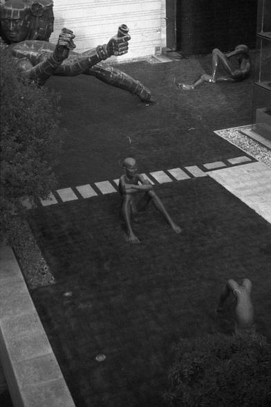 Ilford FP4 Plus 125 (FF)
English Alt Text:
Black and white photo of a garden with four human sculptures in various poses: one lying down with arms raised, one sitting with legs extended, one seated against a wall, and one crouching near a bush. The scene includes grass, paving stones, and a building in the background.

中文替代文字：
黑白照片展示一个花园，四个不同姿势的人体雕塑：一个躺着举起双臂，一个坐着伸直双腿，一个靠墙而坐，一个蹲在灌木旁。场景中有草地、铺石路和背景建筑。