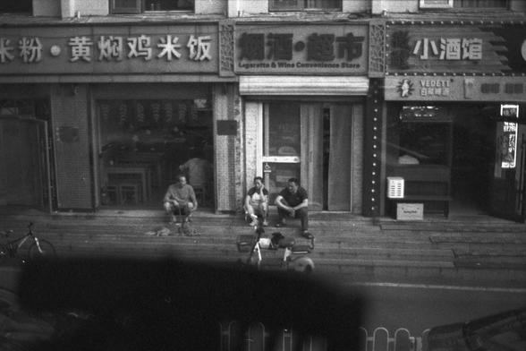 Ilford FP4 Plus 125 (FF)
English Alt Text:
Black and white street scene showing three Chinese storefronts: a restaurant (“Rice noodles, Braised chicken rice”), a convenience store (“Cigarettes & Wine Convenience Store”), and a tavern (“Small Tavern” and “VEDETT White Bear Beer”). Three people sit on the steps chatting, with bicycles parked nearby.

中文替代文字：
黑白街景照片展示三家中国店铺：一家餐馆（“米粉 黄焖鸡米饭”）、一家便利店（“烟酒 超市”）、一家小酒馆（“小酒馆”与“VEDETT 白熊啤酒”）。三人坐在台阶上聊天，旁边停着自行车。