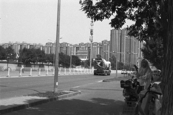 Ilford FP4 Plus 125 (FF)
English Alt Text:
Black and white photo of an urban street scene. A person stands beside a bicycle under a tree in the foreground, holding an object. A cement mixer truck drives down the road, with tall residential buildings and trees in the background. A metal fence lines the street, and a streetlight pole stands on the sidewalk. The image contrasts motion and stillness, nature and urban development.

中文替代文字：
黑白照片展示城市街景。前景中，一人站在树下的自行车旁，手中拿着物品。背景中，一辆混凝土搅拌车在道路上行驶，远处是高层住宅…