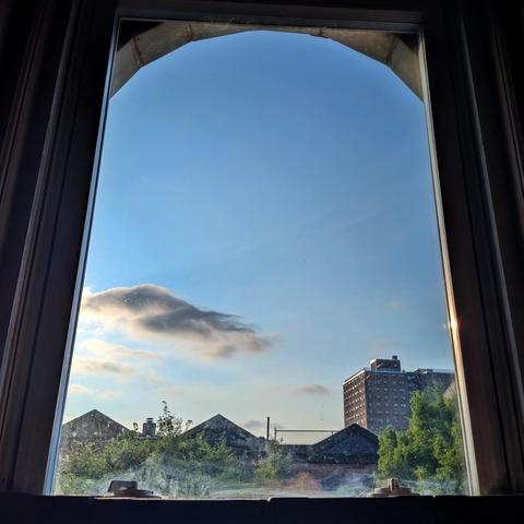 Looking through an arched window seventy-four minutes after sunrise there is one large gray clouds in the mostly clear blue sky. Pointed roofs of Harlem brownstones are silhouetted across the street, and a taller apartment building can be seen in the distance. The green tops of two trees are on the bottom and right. The window is grimy on the bottom. 