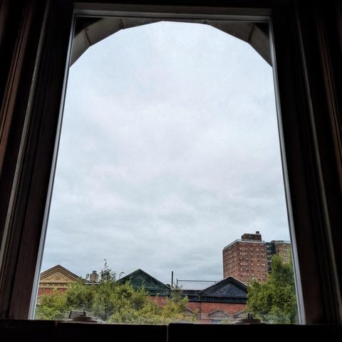 Looking through an arched window two hours after sunrise the sky is filled with light gray clouds. Pointed roofs of Harlem brownstones with red brickwork are across the street, and a taller apartment building can be seen in the distance. The green tops of two trees are on the bottom and right.