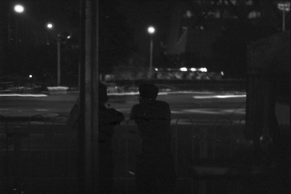 Ilford FP4 Plus 125 (FF)
English Alt Text:
A moody nighttime scene showing two silhouetted figures standing near a reflective glass surface, looking out at blurred streetlights and passing vehicles. Their reflections add depth and duality to the composition.

中文替代文字：
夜晚氛围浓厚的画面，两个人影站在玻璃表面前，望向街道上模糊的灯光和行驶的车辆。他们的倒影增强了画面的层次感和对比。