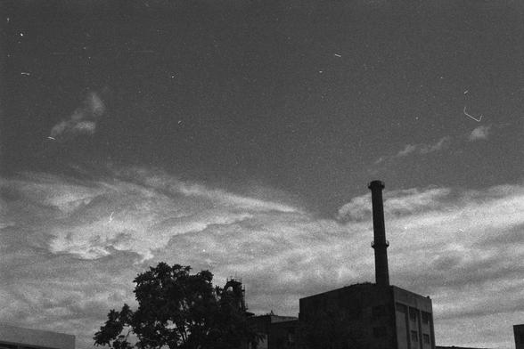 Ilford FP4 Plus 125 (FF)
English Alt Text:
A black and white photograph of an industrial landscape featuring a tall smokestack, a building structure beneath it, and a tree on the left. The cloudy sky and visible scratches suggest the photo is aged or taken with film.

中文替代文字：
一张黑白工业景观照片，画面右侧是一座高大的烟囱，下方是建筑结构，左侧有一棵树。天空布满云层，照片上有划痕，显示其年代久远或使用胶片拍摄。