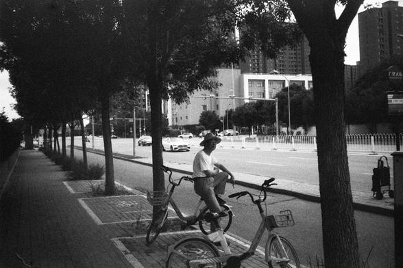 English Alt Text:
A black and white photo of a quiet urban street. A person wearing a hat and light clothing sits on a bicycle parked near a tree on the sidewalk. Another bicycle is nearby. Trees line the left side of the street, while cars and buildings fill the background, creating a calm and reflective atmosphere.

中文替代文字：
一张黑白照片，描绘一条宁静的城市街道。一位戴帽子、穿浅色衣服的人坐在人行道树旁停放的自行车上，旁边还有另一辆自行车。街道左侧种有树木，背景中有汽车和建筑，营造出平静而富有思考感的氛围。