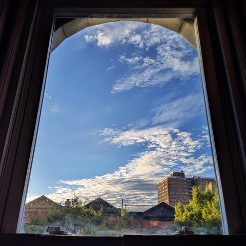 Looking through an arched window an hour after sunrise the bright blue sky is half-filled with swirling white clouds. Pointed roofs of Harlem brownstones are silhouetted across the street, and a taller apartment building can be seen in the distance. The green tops of two trees are on the bottom and right.