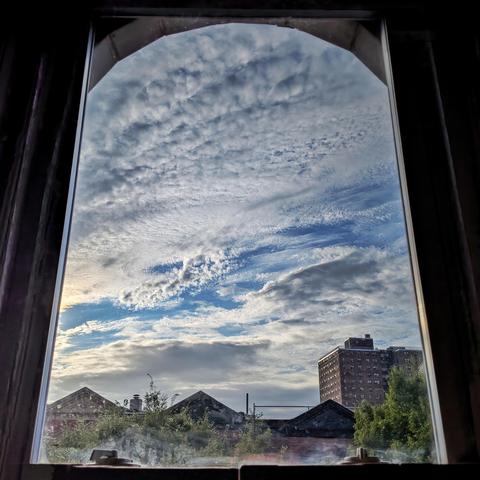 Looking through an arched window ninety minutes after sunrise the blue sky is filled with complex windswept white clouds. Pointed roofs of Harlem brownstones are silhouetted across the street, and a taller apartment building can be seen in the distance. The green tops of two trees are on the bottom and right.