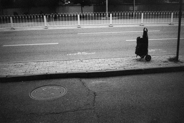 English Alt Text:
A black and white photo of a wheeled shopping cart placed on a brick sidewalk near a road. The road has white lane markings and a white fence. A manhole cover is visible on the sidewalk, and the numbers “1918” and “1919” are painted on the road in Chinese characters.

中文替代文字：
一张黑白照片，画面中一个带轮子的购物车停在人行道上，靠近道路。道路上有白色车道线和白色护栏。人行道上有一个井盖，路面上用中文标注了“1918”和“1919”的数字。