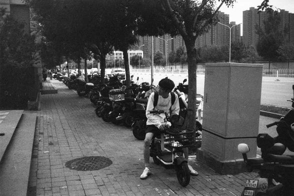 English Alt Text:
A black and white photo of a person sitting on a scooter parked on a brick-paved sidewalk lined with other scooters and motorcycles. The person wears a long-sleeved shirt, shorts, sneakers, and a backpack. Trees line the sidewalk, and tall buildings and cars are visible in the background.

中文替代文字：
一张黑白照片，画面中一位穿长袖衬衫、短裤、运动鞋并背着背包的人坐在一辆停在人行道上的电动车上。人行道铺有砖块，两旁停满了摩托车和电动车，边缘种有树木。背景中可见高楼和行驶的汽车。