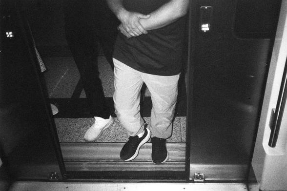 English Alt Text:
Black-and-white photo of two people standing in the doorway of a subway car. One person wears light pants and dark sneakers, standing casually with legs crossed. The other wears dark pants and white shoes. The train doors are open, revealing the platform.

中文替代文字：
一张黑白照片，两人站在地铁车门口。一人穿浅色裤子和深色运动鞋，交叉站立，姿态随意；另一人穿深色裤子和白鞋。车门敞开，地铁站台清晰可见。