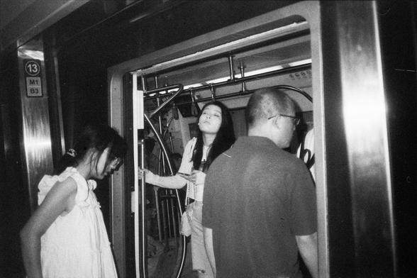 English Alt Text:
Black-and-white photo of three people at the entrance of a subway train. One person in a white dress looks down, another stares directly at the camera while holding a pole, and a third faces away. Subway interior and signage with “13” and “M1 B3” are visible.

中文替代文字：
一张黑白照片，三人站在地铁车门口。一人穿白裙低头看地，另一人握着扶杆直视镜头，第三人背对镜头。车厢内部和标有“13”和“M1 B3”的标识清晰可见。