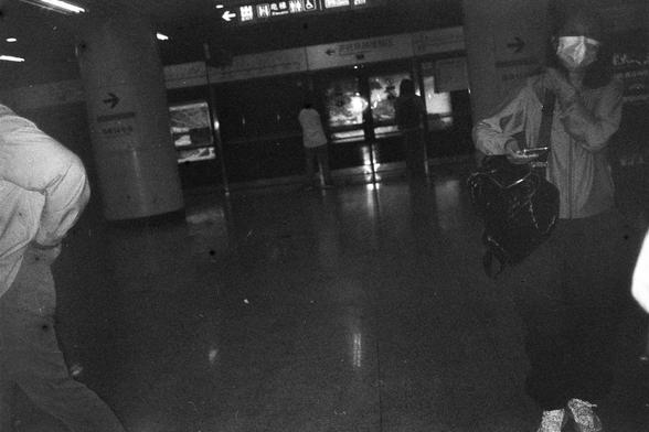 English Alt Text:
A black-and-white photo of a subway station interior. Several people are present, including one wearing a face mask and holding a phone with a large bag over their shoulder. Directional signs with Chinese characters and arrows are visible on columns, and illuminated signage hangs above a counter area in the background.

中文替代文字：
一张地铁站内部的黑白照片。画面中有几位乘客，其中一人戴口罩、手持手机，肩背大包。柱子上有带箭头的中文方向指示牌，背景中柜台上方悬挂着发光的中文标识牌，可能是车站信息。