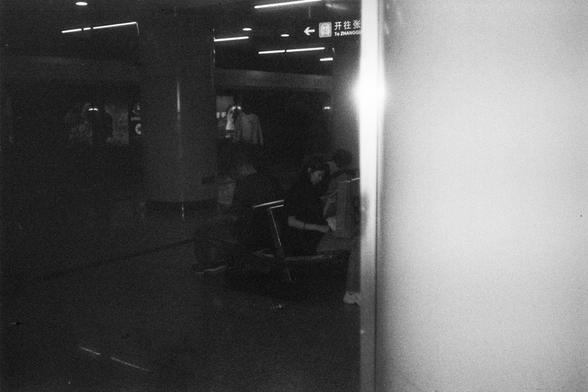English Alt Text:
A grainy black-and-white photo of a dimly lit subway station with people sitting on benches. Large columns and overhead lights frame the space. A sign with Chinese characters and an arrow points left, indicating train departure information.

中文替代文字：
一张昏暗地铁站的黑白照片，几位乘客坐在长椅上。画面中有粗大的柱子和顶部照明灯。中央上方有一个写着“开往 班次”的中文指示牌，箭头指向左侧，提示列车发车信息。