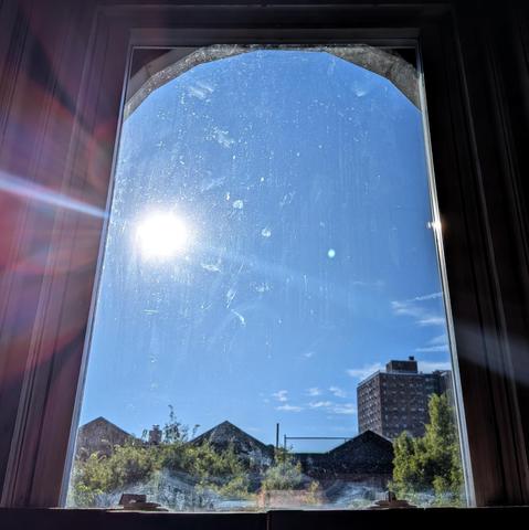 Looking through an arched window two hours after sunrise the sun is above the buildings in the blue sky, making a dramatic lens flare with multi-colored petals on the left. Pointed roofs of Harlem brownstones are silhouetted across the street, and a taller apartment building can be seen in the distance. The green tops of two trees are on the bottom and right.