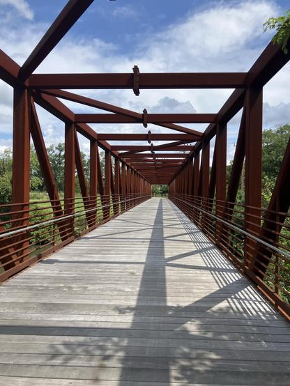 A view of a pedestrian bridge with a rust-colored steel frame, wooden planks, and green foliage on either side. The sky is partly cloudy, and shadows cast by the structure create interesting patterns on the walkway.